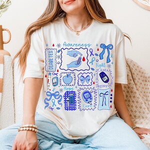 May include: Cream-colored t-shirt with blue and white graphics related to diabetes awareness. The design includes the words "Awareness", "Diabetes", "Hope", and "Cure", along with illustrations of a blood glucose meter, insulin pen, and a blue ribbon.