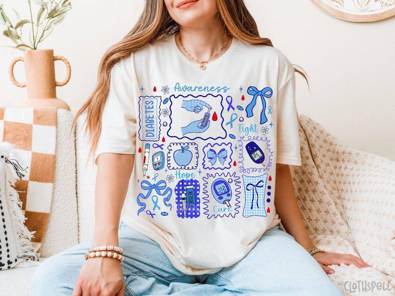 May include: Cream-colored t-shirt with blue and white graphics related to diabetes awareness. The design includes the words "Awareness", "Diabetes", "Hope", and "Cure", along with illustrations of a blood glucose meter, insulin pen, and a blue ribbon.