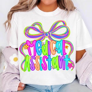 Puede incluir: Camiseta blanca con la frase "MEDICAL ASSISTANT" en letras coloridas. El dise&ntilde;o incluye un gran lazo multicolor y elementos decorativos de flores y estrellas.