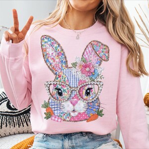 May include: Pink sweatshirt featuring a whimsical bunny design. The bunny has floral patterned ears, a patchwork face, and wears rhinestone-studded glasses. It is adorned with flowers and carrots, perfect for Easter or spring. The sweatshirt is a soft pink color.