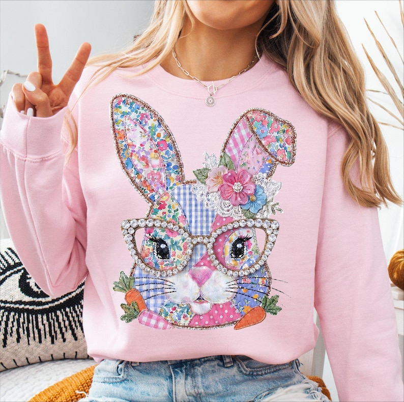 May include: Pink sweatshirt featuring a whimsical bunny design. The bunny has floral patterned ears, a patchwork face, and wears rhinestone-studded glasses. It is adorned with flowers and carrots, perfect for Easter or spring. The sweatshirt is a soft pink color.