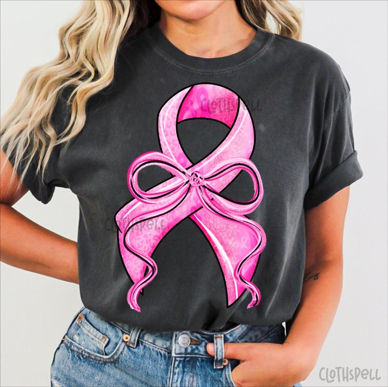 May include: Dark gray t-shirt featuring a large pink ribbon design with a bow, outlined in black. The ribbon has a textured pattern and the word "Clothspell" is visible. The shirt is a casual style.