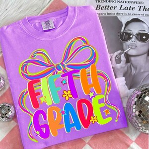 May include: A lilac-colored t-shirt with a colorful bow and the words "FIFTH GRADE" in bright, playful letters. The design includes stars and flowers. The shirt is a Comfort Colors brand.