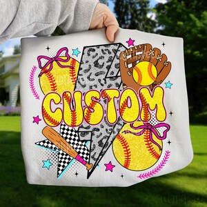 May include: White tote bag featuring a softball theme. The design includes yellow softballs, a baseball glove, a bat, and the word "CUSTOM" in yellow bubble letters. Accents include pink bows, stars, and a lightning bolt.