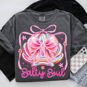 K&ouml;nnte beinhalten: Dunkelgraues Sweatshirt mit einem bunten Muscheldesign, einer gro&szlig;en rosa Schleife, Perlenakzenten und dem Schriftzug "Salty Soul". Das Design umfasst rosa Sterne und Blumen. Ein Handy mit karierter H&uuml;lle liegt rechts.