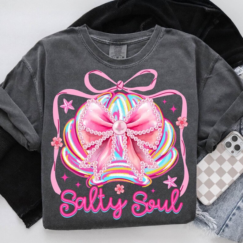 K&ouml;nnte beinhalten: Dunkelgraues Sweatshirt mit einem bunten Muscheldesign, einer gro&szlig;en rosa Schleife, Perlenakzenten und dem Schriftzug "Salty Soul". Das Design umfasst rosa Sterne und Blumen. Ein Handy mit karierter H&uuml;lle liegt rechts.