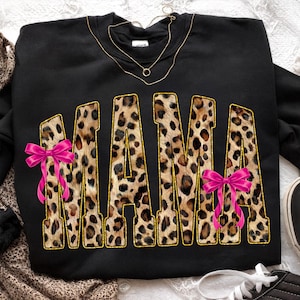 May include: Black sweatshirt with the word "MAMA" in leopard print letters outlined in gold. Pink bows are on the left and right side of the word. A gold necklace is draped across the top of the sweatshirt.