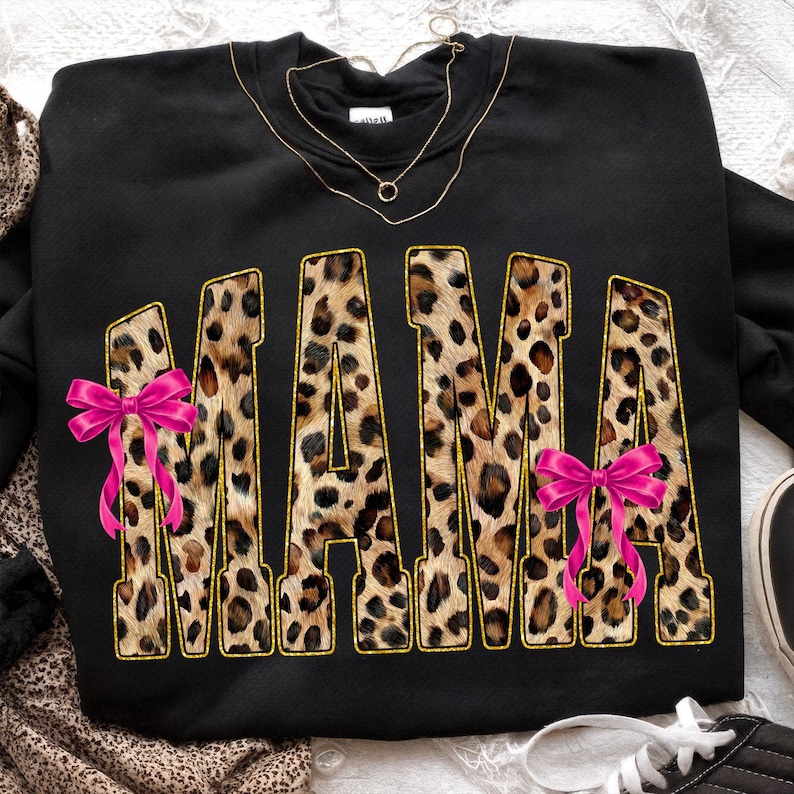 May include: Black sweatshirt with the word "MAMA" in leopard print letters outlined in gold. Pink bows are on the left and right side of the word. A gold necklace is draped across the top of the sweatshirt.