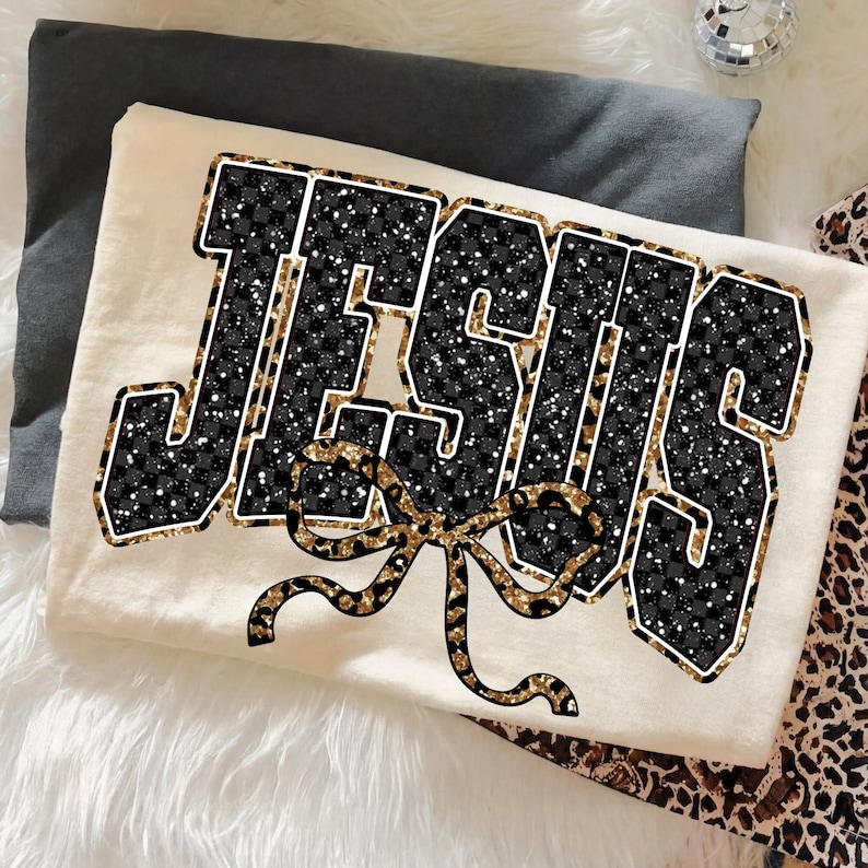 May include: Cream-colored t-shirt featuring the word "JESUS" in a bold, black and gold leopard print design. A gold leopard print bow is centered below the word. The shirt is folded on a white fluffy surface.