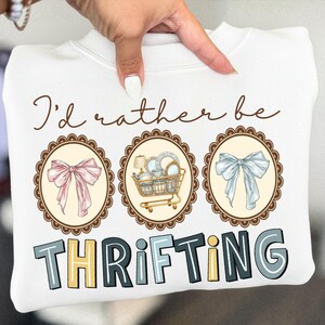 May include: White sweatshirt with the text "I'd rather be thrifting." The design includes three oval frames with a pink bow, a shopping cart with items, and a blue bow. The word "THRIFTING" is in block letters.
