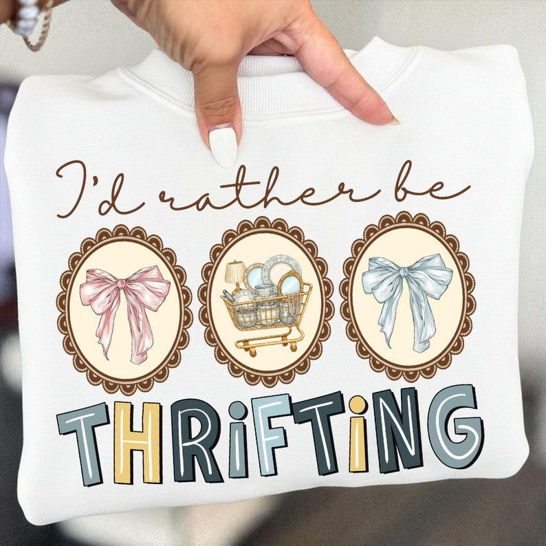 May include: White sweatshirt with the text "I'd rather be thrifting." The design includes three oval frames with a pink bow, a shopping cart with items, and a blue bow. The word "THRIFTING" is in block letters.