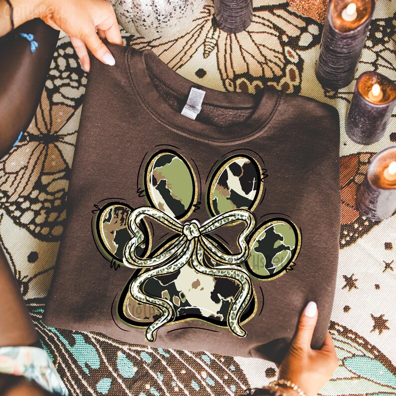 K&ouml;nnte beinhalten: Ein braunes Sweatshirt mit einem Pfotenabdruck-Design und einer Schleife. Der Pfotenabdruck hat ein Camouflage-Muster in Gr&uuml;n-, Braun- und Wei&szlig;t&ouml;nen. Die Schleife ist wei&szlig; mit goldenem Umriss. Das Sweatshirt wird von H&auml;nden gehalten.