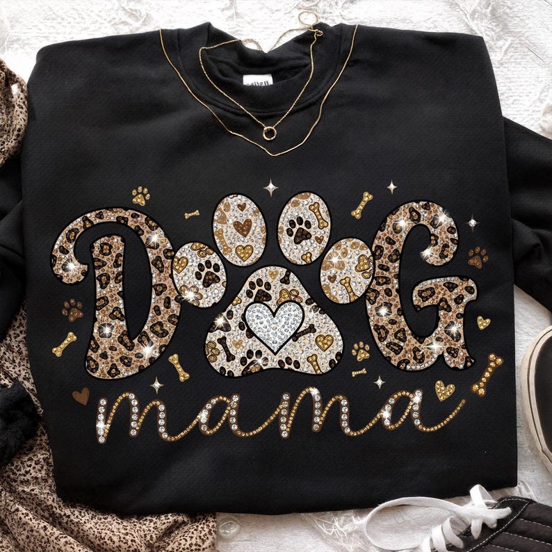 May include: Black sweatshirt with the words "DOG MAMA" in a sparkly, leopard print design. The paw print in the center has a heart. Gold necklace is draped over the sweatshirt.