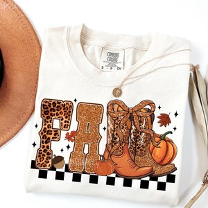 Peut inclure: T-shirt cr&egrave;me avec un motif "FALL". Les lettres sont d&eacute;cor&eacute;es d'imprim&eacute; l&eacute;opard et de paillettes. Le motif comprend des bottes de cowboy, des citrouilles et un gland. Un collier dor&eacute; et un chapeau marron sont &eacute;galement visibles.