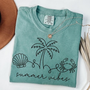 May include: A seafoam green t-shirt with a black graphic of a seashell, palm tree, and crab, with the words "summer vibes" in a script font. A gold necklace with a pendant is on the shirt.