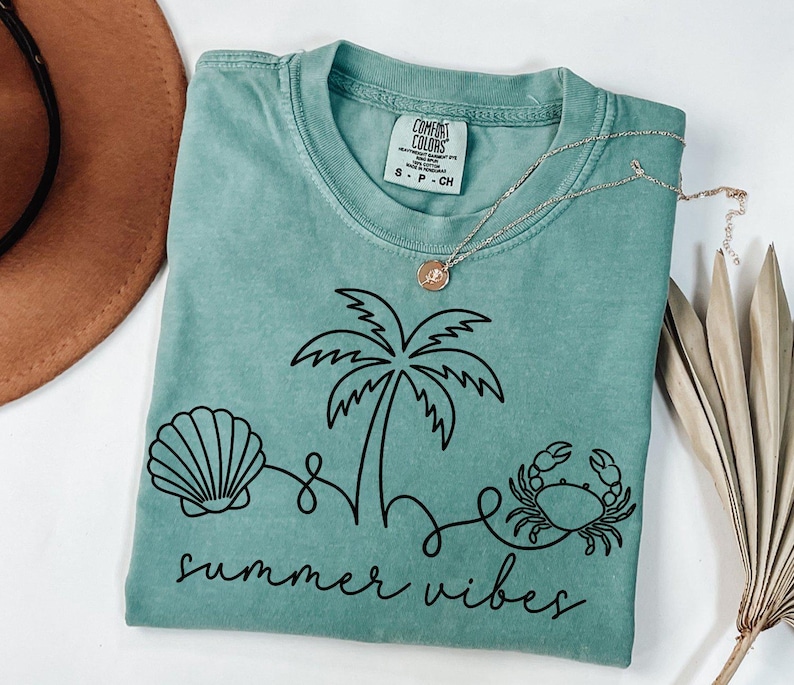 May include: A seafoam green t-shirt with a black graphic of a seashell, palm tree, and crab, with the words "summer vibes" in a script font. A gold necklace with a pendant is on the shirt.