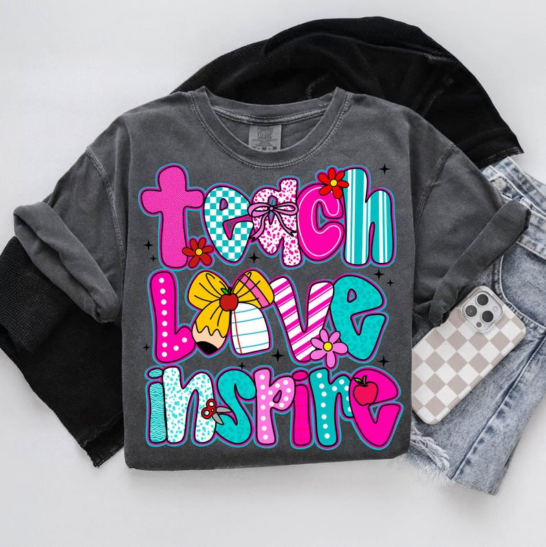 May include: Dark gray long-sleeve t-shirt with the words "Teach Love Inspire" in colorful bubble letters. The design includes flowers, bows, and school-themed graphics. The shirt is paired with denim shorts and a phone.