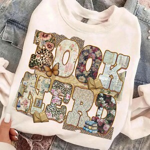 May include: White crew neck sweatshirt with the words "BOOK NERD" in a colorful design. The letters are filled with floral, leopard print, and book page patterns. A butterfly and small books are also featured.