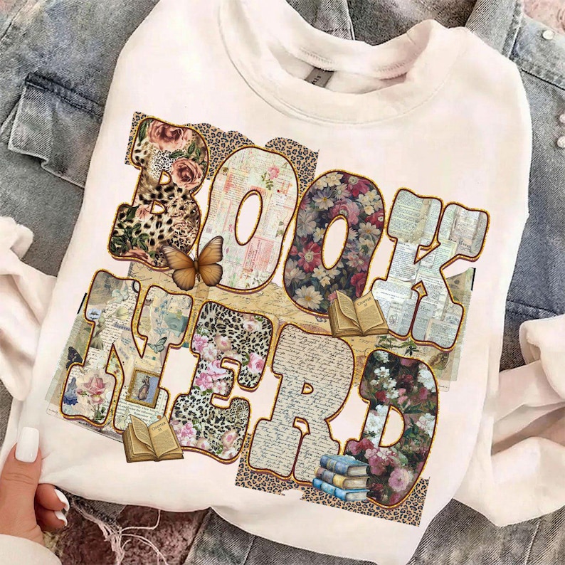 May include: White crew neck sweatshirt with the words "BOOK NERD" in a colorful design. The letters are filled with floral, leopard print, and book page patterns. A butterfly and small books are also featured.