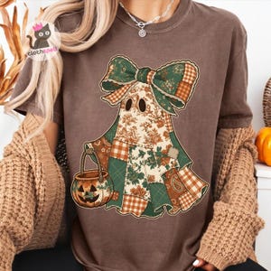 May include: A brown short-sleeve t-shirt featuring a graphic of a ghost wearing a large green and brown patchwork bow. The ghost is composed of various floral and plaid fabric patterns and is holding a matching patchwork pumpkin candy bucket.