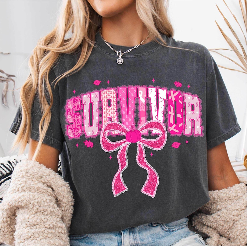 May include: Dark gray t-shirt with the word "SURVIVOR" in pink letters, decorated with pink ribbons and a large pink bow. The shirt is a casual style, suitable for everyday wear.