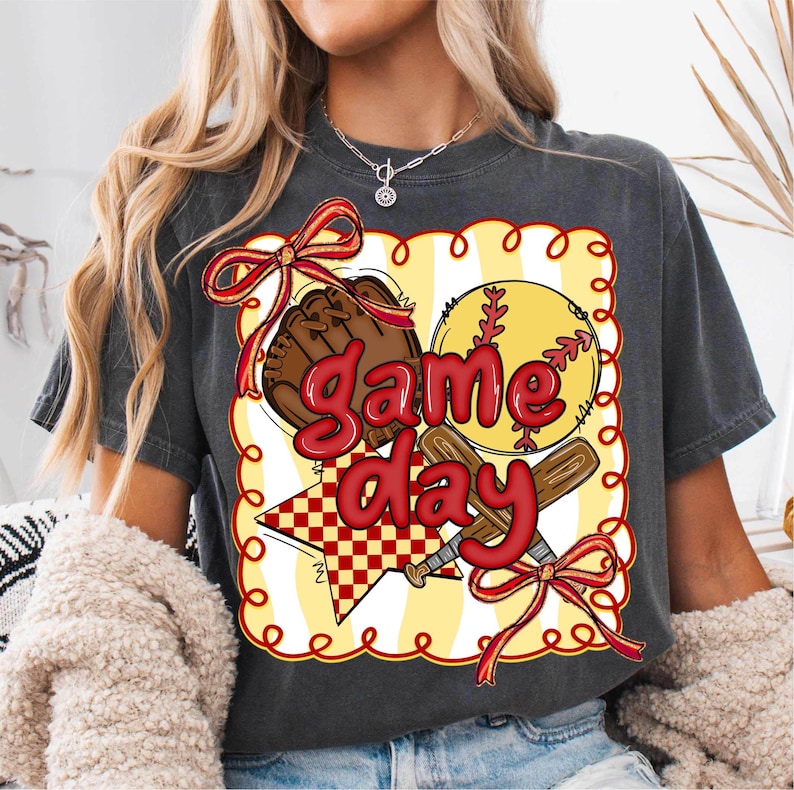 Puede incluir: Camiseta gris oscuro con un dise&ntilde;o de b&eacute;isbol. El dise&ntilde;o incluye una pelota de b&eacute;isbol, un guante y un bate, con las palabras "Game Day" en rojo. El dise&ntilde;o est&aacute; enmarcado con un fondo a rayas amarillas y blancas y lazos rojos.