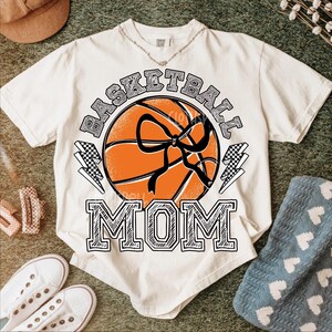 Pode incluir: Camiseta creme com um gr&aacute;fico de basquete e as palavras "BASKETBALL MOM". A bola de basquete &eacute; laranja com detalhes em preto e um la&ccedil;o preto. Dois raios com um padr&atilde;o quadriculado est&atilde;o em ambos os lados da bola.