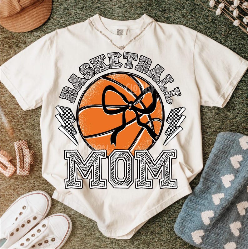 Pode incluir: Camiseta creme com um gr&aacute;fico de basquete e as palavras "BASKETBALL MOM". A bola de basquete &eacute; laranja com detalhes em preto e um la&ccedil;o preto. Dois raios com um padr&atilde;o quadriculado est&atilde;o em ambos os lados da bola.