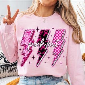 May include: A light pink sweatshirt featuring three lightning bolt graphics. Each bolt has a different design: pink ribbons, black ribbons, and a pink and white checkerboard pattern. The words "pink out" are written across the bolts.
