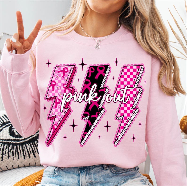 May include: A light pink sweatshirt featuring three lightning bolt graphics. Each bolt has a different design: pink ribbons, black ribbons, and a pink and white checkerboard pattern. The words "pink out" are written across the bolts.