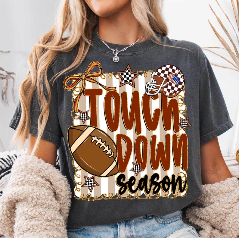 May include: Dark gray t-shirt with a football-themed design. The design includes a football, a helmet, stars, and the words "Touch Down Season" in brown and black. The background has vertical stripes.