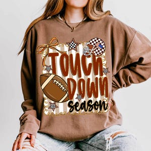 May include: Brown sweatshirt with the words "Touch Down Season" in a bold, decorative font. The design includes a football, a helmet with an American flag design, and stars. The sweatshirt has a crew neck and long sleeves.