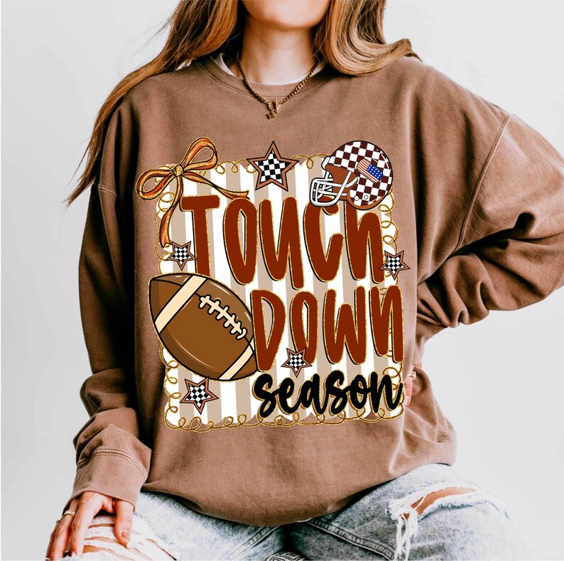 May include: Brown sweatshirt with the words "Touch Down Season" in a bold, decorative font. The design includes a football, a helmet with an American flag design, and stars. The sweatshirt has a crew neck and long sleeves.