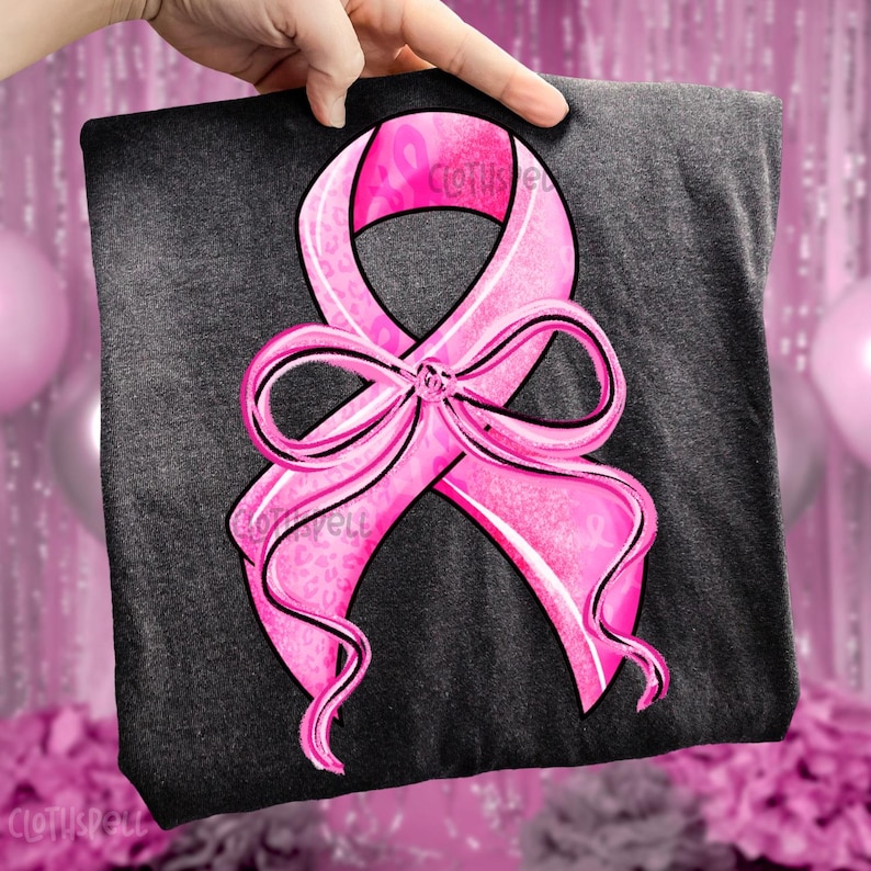 May include: A dark gray t-shirt featuring a large pink ribbon design with a bow. The ribbon has a black outline and a leopard print pattern. The text "Clothspell" is visible on the shirt.