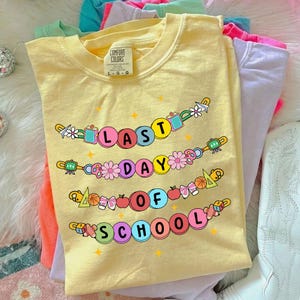 May include: Yellow Comfort Colors t-shirt with the text "LAST DAY OF SCHOOL" in colorful bubble letters. The design includes flowers, school supplies, and stars. Other pastel colored t-shirts are visible in the background.
