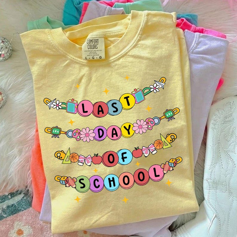 May include: Yellow Comfort Colors t-shirt with the text "LAST DAY OF SCHOOL" in colorful bubble letters. The design includes flowers, school supplies, and stars. Other pastel colored t-shirts are visible in the background.