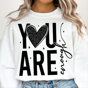 Peut inclure: Sweat-shirt blanc avec un texte noir qui dit "YOU ARE enough" avec un c&oelig;ur noir imprim&eacute; l&eacute;opard au milieu.