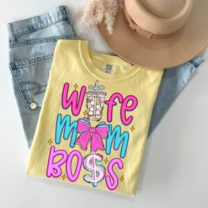 May include: A yellow t-shirt with the words "Wife Mom Boss" in pink and blue, along with a graphic of a drink and a pink bow. The shirt is laid on top of a pair of blue jeans, a tan hat, and a necklace.