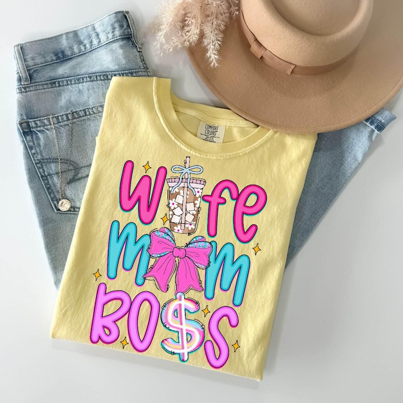 May include: A yellow t-shirt with the words "Wife Mom Boss" in pink and blue, along with a graphic of a drink and a pink bow. The shirt is laid on top of a pair of blue jeans, a tan hat, and a necklace.