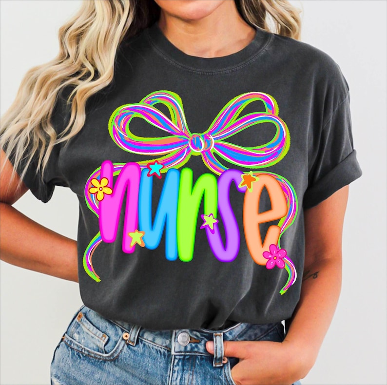 May include: Dark grey t-shirt featuring the word "NURSE" in vibrant colours. The word is decorated with stars and flowers, and topped with a large, multi-coloured bow. The t-shirt is a casual style, suitable for everyday wear.