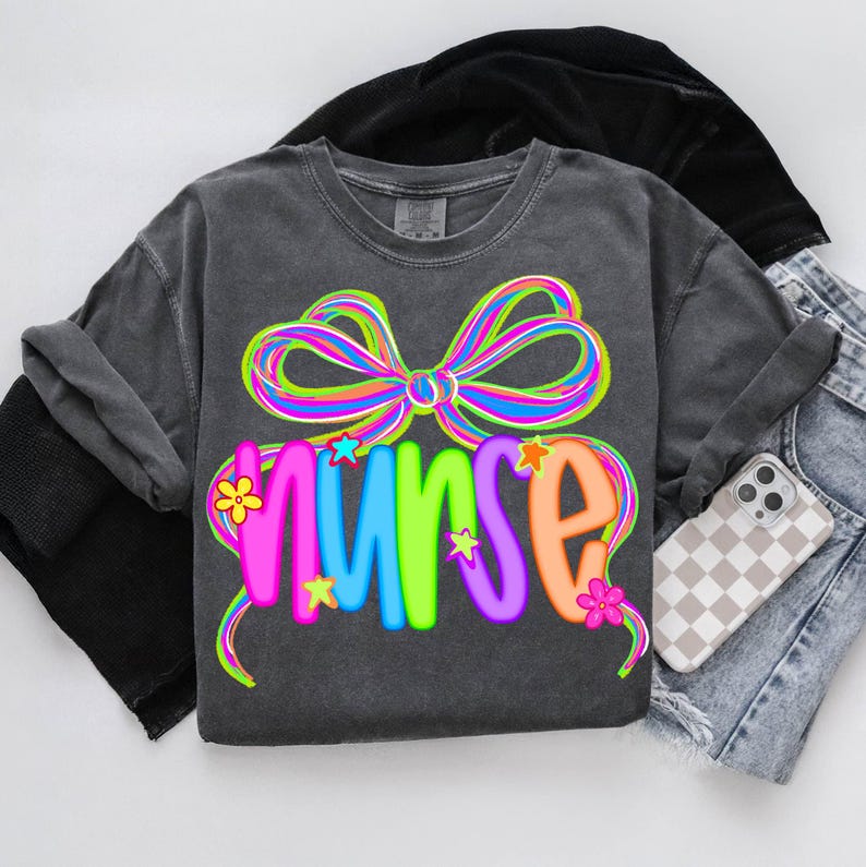 May include: Charcoal grey t-shirt featuring a vibrant, multi-coloured bow design above the word "nurse". The word is in bright colours with star and flower accents. The shirt is styled with denim shorts and a phone.