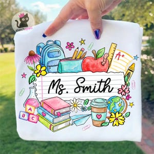 Pode incluir: Camisola branca com um design colorido com tema escolar. O design inclui uma mochila, lápis, uma maçã, livros, um globo e o nome "Ms. Smith" num fundo de papel pautado. O design é rodeado por flores e estrelas.