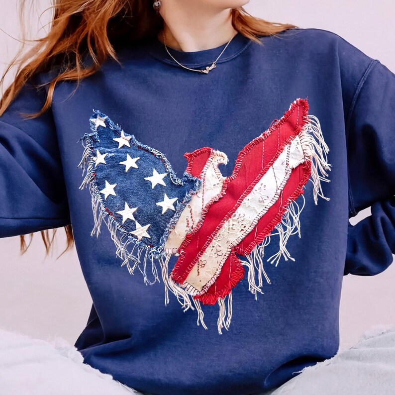 May include: Navy blue sweatshirt featuring a patriotic eagle design. The eagle is crafted from denim with white stars, red and white striped fabric, and white lace accents. The design has a distressed, fringed edge.