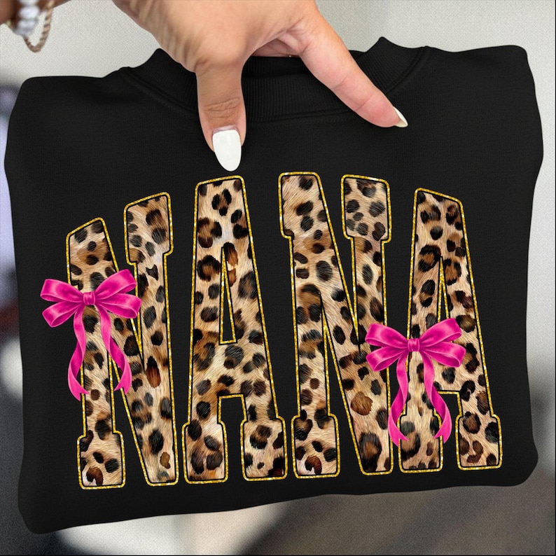 May include: Black sweatshirt with the word "NANA" in leopard print letters outlined in gold. Pink bows are attached to the letters "N" and "A".