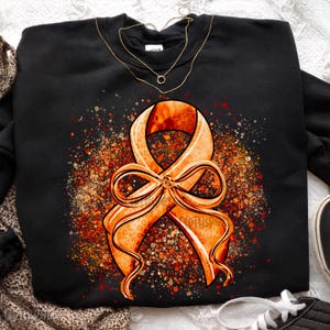 May include: Black sweatshirt featuring a large orange ribbon with a bow, surrounded by a speckled design in shades of orange and brown. A gold necklace is visible at the neckline. The ribbon design has a leopard print pattern.