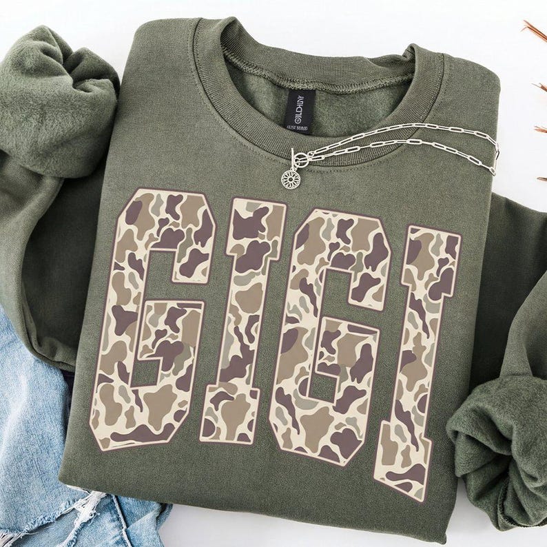 May include: An olive green sweatshirt with the word "GIGI" in large, block letters. The letters have a camouflage pattern in shades of brown and tan. A silver necklace with a pendant is draped on the sweatshirt.