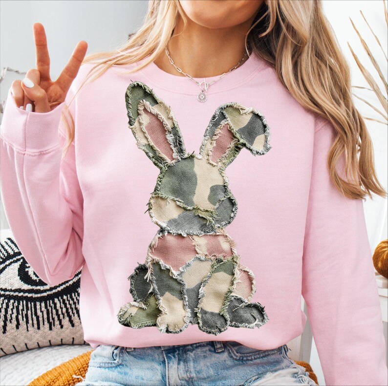 May include: A light pink sweatshirt featuring a patchwork bunny design in shades of green, pink, and tan. The bunny is centered on the front of the long-sleeved top. The sweatshirt has a crew neck and a relaxed fit.