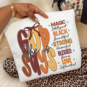 May include: White sweatshirt with the text "GOD SAYS She is" in brown, orange, and yellow. Additional words include "MAGIC, BLACK, STRONG, BLESSED, LOVE" and other positive affirmations. A brown bow is at the top. The sweatshirt is held by a person.