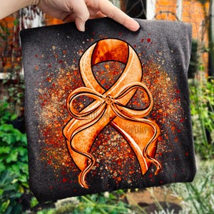 May include: A dark gray t-shirt featuring a large orange awareness ribbon with a bow. The ribbon has a leopard print pattern and is surrounded by orange and red speckles. The shirt is held up by a hand.