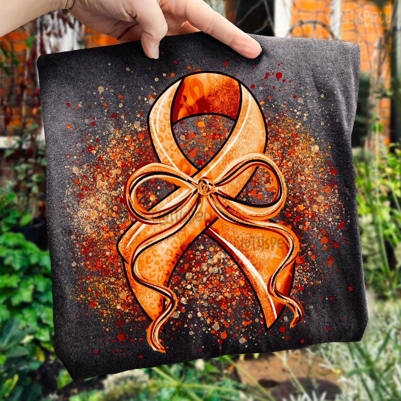 May include: A dark gray t-shirt featuring a large orange awareness ribbon with a bow. The ribbon has a leopard print pattern and is surrounded by orange and red speckles. The shirt is held up by a hand.